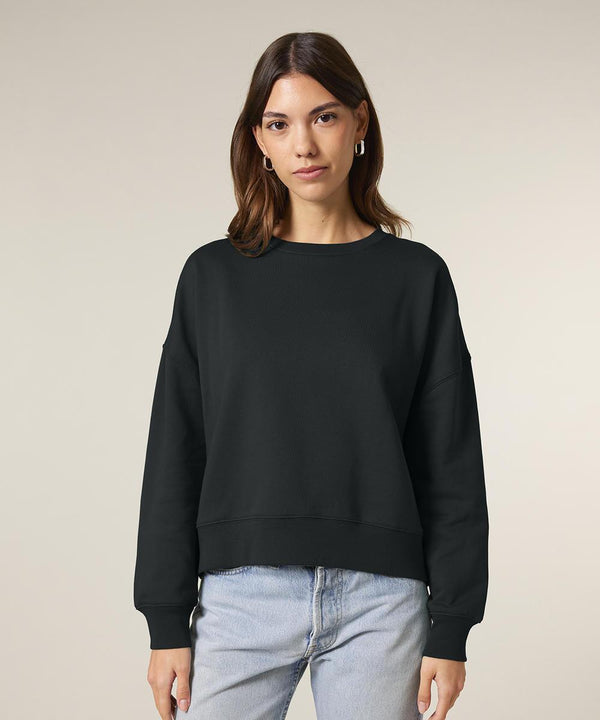 A woman with straight brown hair wears the black Stanley/Stella Stella Alma sweatshirt (STSW212) and light blue jeans, standing facing the camera against a plain, light-coloured background with a neutral expression.