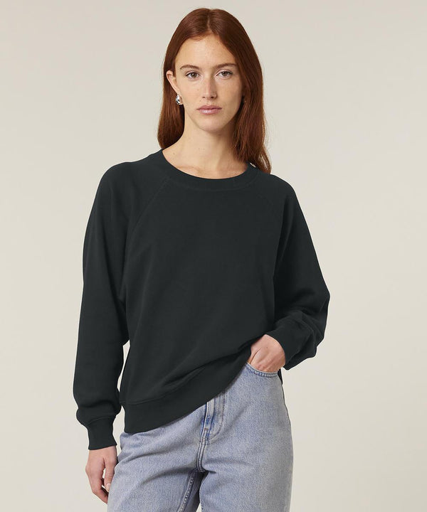 A person with long red hair is wearing the Stanley/Stella Stella Clara (STSW217) black sweatshirt and light blue jeans, standing against a neutral background with one hand in their pocket.