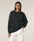 A person with long dark hair wears the Stanley/Stella Stella Paloma (STSW218) black sweatshirt and white cargo trousers, standing against a plain light background with one hand in a pocket.