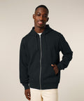 A person faces the camera, wearing the Stanley/Stella Mixer (STSU205) black zip-up hoodie with their left hand in the pocket and beige trousers, set against a plain light-coloured background.