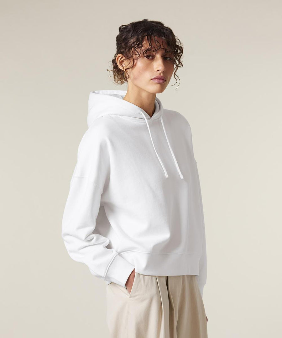 A person with curly hair wears the Stanley/Stella Stella Nora (STSW213) white hoodie with the hood down and beige trousers, standing against a light background with hands in pockets, facing slightly to the side.