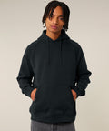 A person with medium-length dreadlocks faces the camera, hands in the front pocket of a black Stanley/Stella Striker (STSU202) hooded sweatshirt. The background is plain and light-coloured.