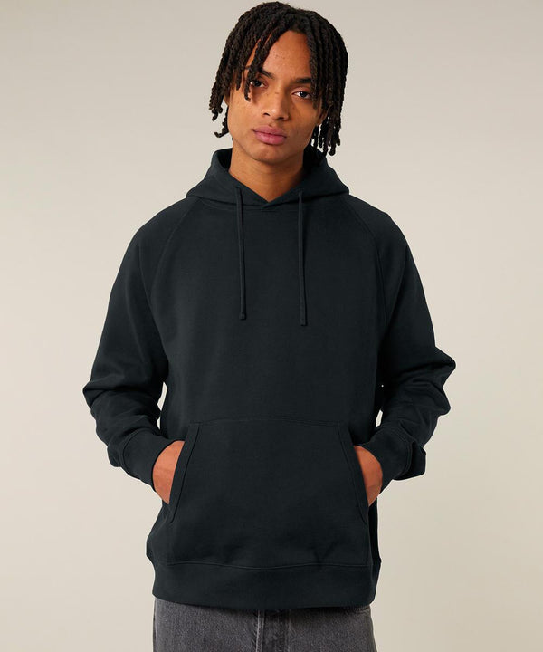 A person with medium-length dreadlocks faces the camera, hands in the front pocket of a black Stanley/Stella Striker (STSU202) hooded sweatshirt. The background is plain and light-coloured.