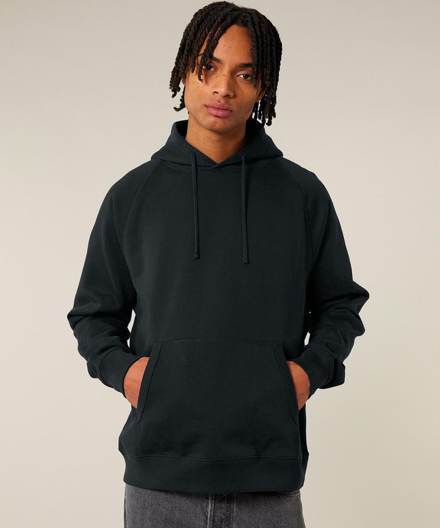 A person with medium-length dreadlocks faces the camera, hands in the front pocket of a black Stanley/Stella Striker (STSU202) hooded sweatshirt. The background is plain and light-coloured.