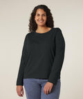 A person with curly hair is standing and smiling, wearing the Stanley/Stella Stella Muser long sleeve (STTW219) and blue jeans against a plain light background.
