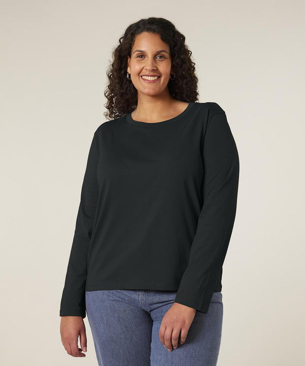 A person with curly hair is standing and smiling, wearing the Stanley/Stella Stella Muser long sleeve (STTW219) and blue jeans against a plain light background.