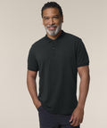 A smiling man with short curly hair and a salt-and-pepper beard stands against a plain light background, wearing the Stanley/Stella Stanley Coaster (STPM224) black short-sleeve polo shirt with one hand in his pocket.