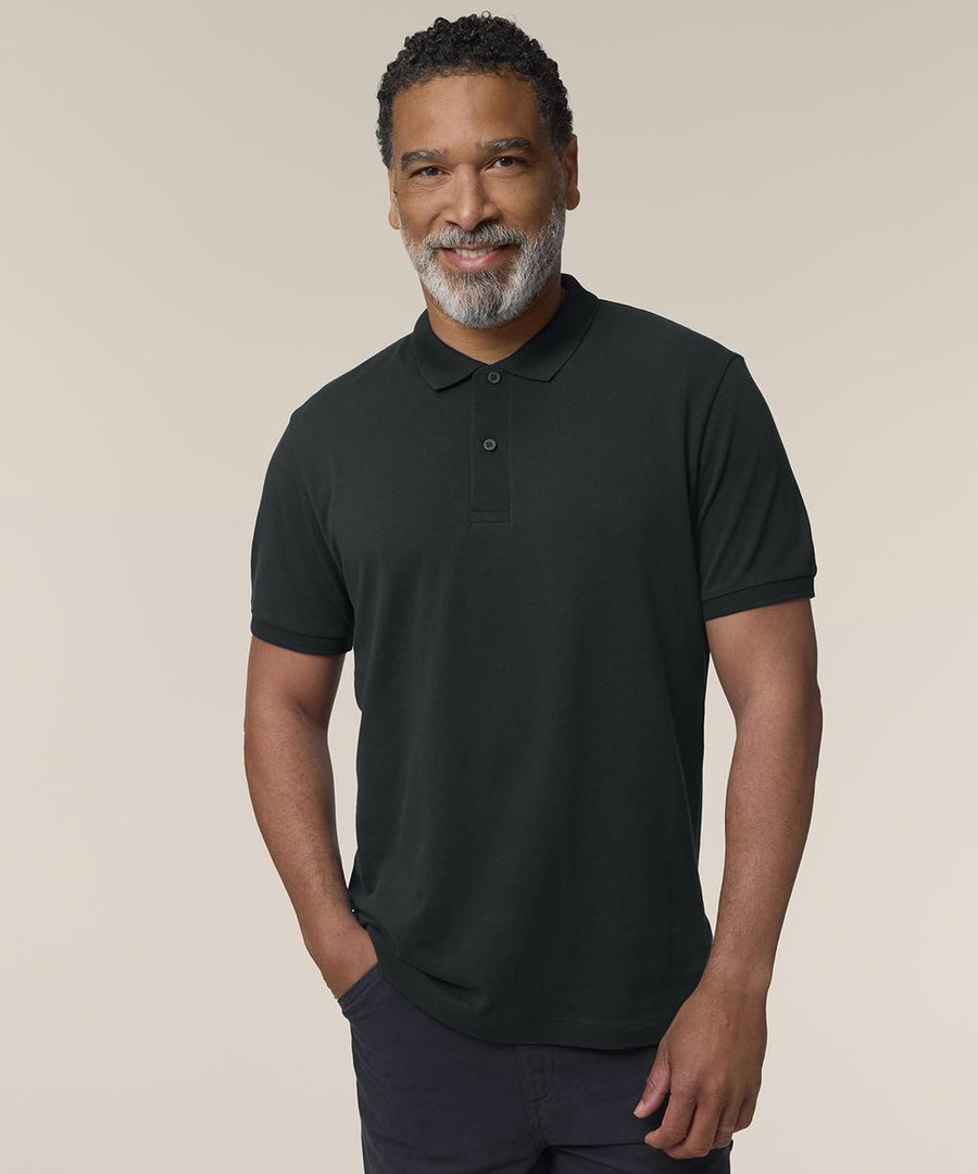 A smiling man with short curly hair and a salt-and-pepper beard stands against a plain light background, wearing the Stanley/Stella Stanley Coaster (STPM224) black short-sleeve polo shirt with one hand in his pocket.