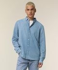 A person with short, bleached blond hair wears the Stanley/Stella Stanley denim shirt (STWM967) over a white T-shirt and light blue jeans, standing against a plain, light-coloured background.