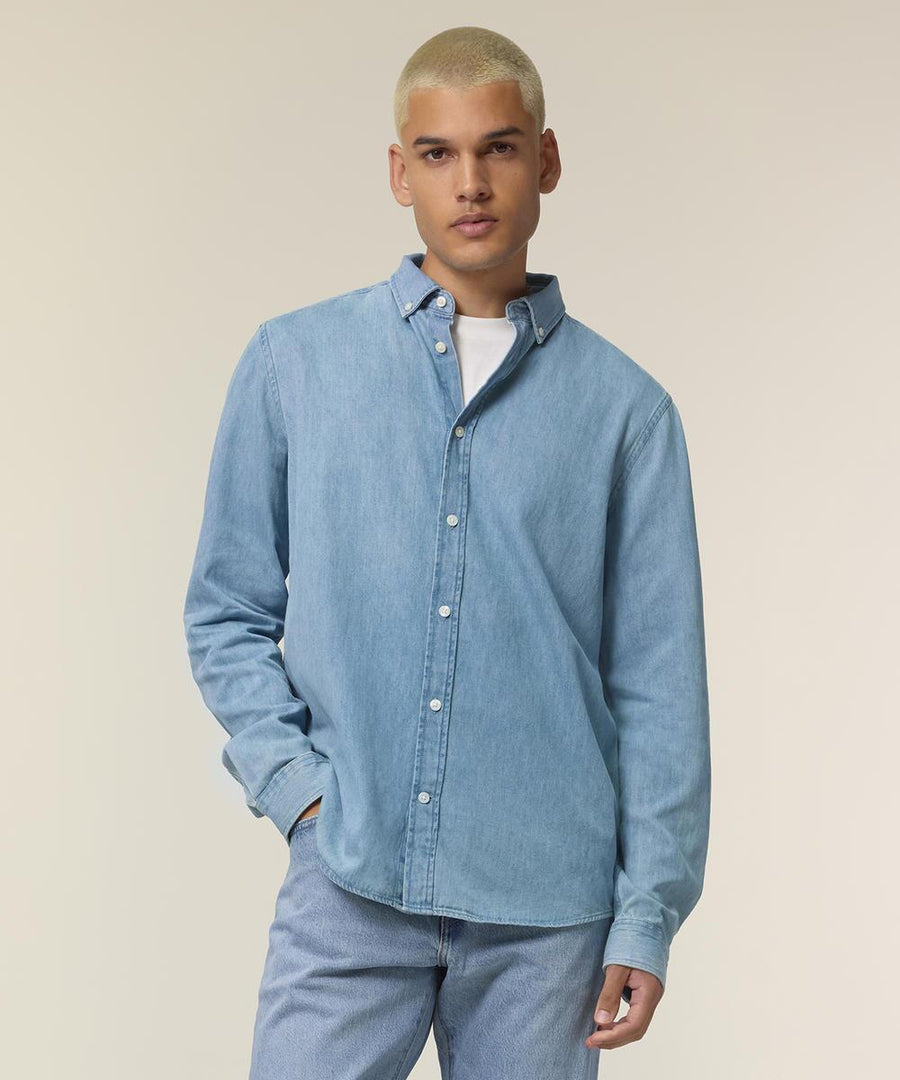 A person with short, bleached blond hair wears the Stanley/Stella Stanley denim shirt (STWM967) over a white T-shirt and light blue jeans, standing against a plain, light-coloured background.