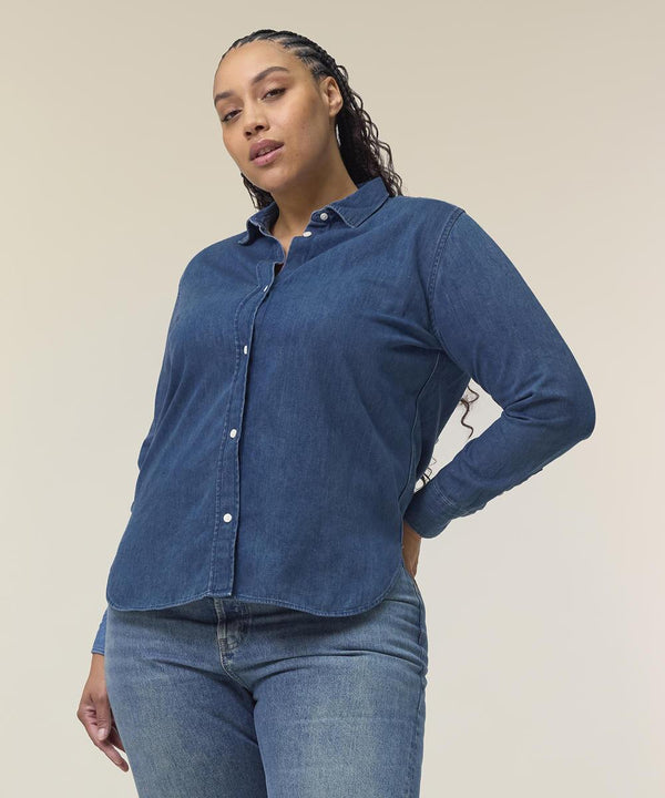 A person with long, braided hair wears the Stanley/Stella Stella denim shirt (STWW970) and blue jeans, standing against a plain light background with one hand on their hip.