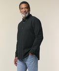 A man with short curly hair and a beard wears the Stanley/Stella Stanley Oxford shirt (STWM968) in black and light blue jeans. He stands against a plain, light background, looking off to the side with a slight smile.