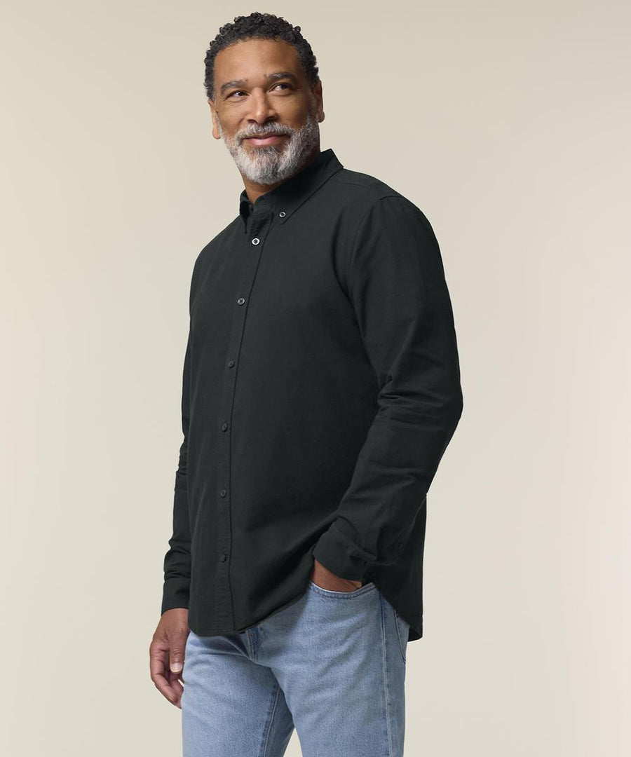 A man with short curly hair and a beard wears the Stanley/Stella Stanley Oxford shirt (STWM968) in black and light blue jeans. He stands against a plain, light background, looking off to the side with a slight smile.