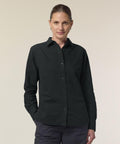 A person with light skin and brown hair tied back stands against a plain background, wearing the black Stanley/Stella Stella Oxford shirt (STWW971) and dark trousers, with one hand in their pocket.