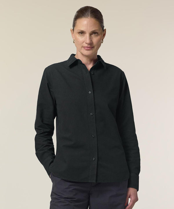 A person with light skin and brown hair tied back stands against a plain background, wearing the black Stanley/Stella Stella Oxford shirt (STWW971) and dark trousers, with one hand in their pocket.