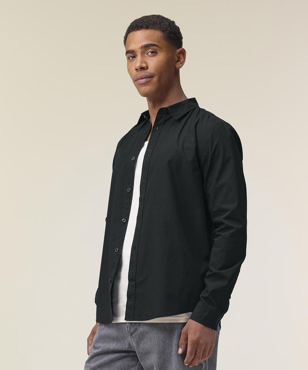 A man stands slightly turned, wearing the Stanley/Stella Stanley Styler shirt (STWM969) in black over a white T-shirt with grey trousers. The background is plain and light-coloured.