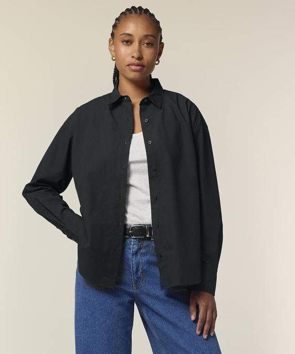 A person with plaited hair wears the Stanley/Stella Stella Styler shirt (STWW973) in black over a white top, paired with blue jeans and a black belt, standing against a plain light background with one hand on their hip.