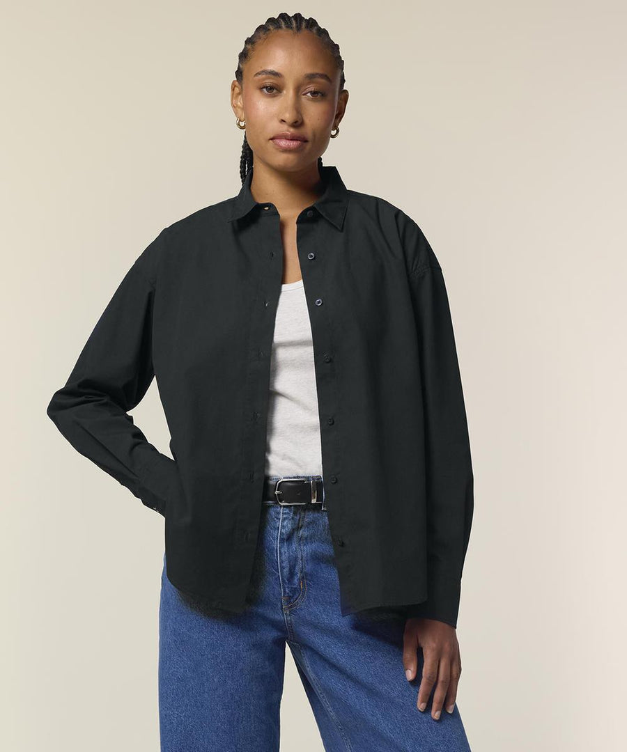 A person with plaited hair wears the Stanley/Stella Stella Styler shirt (STWW973) in black over a white top, paired with blue jeans and a black belt, standing against a plain light background with one hand on their hip.