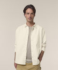 A man with medium-length brown hair wears the Stanley/Stella Worker shirt (STWU975), cream-coloured, over a beige t-shirt and tan trousers. He stands against a plain light background, one hand in his pocket, looking at the camera.