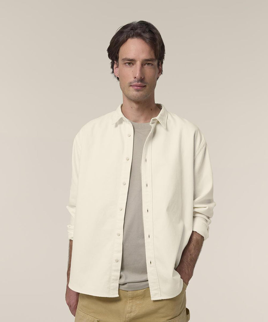 A man with medium-length brown hair wears the Stanley/Stella Worker shirt (STWU975), cream-coloured, over a beige t-shirt and tan trousers. He stands against a plain light background, one hand in his pocket, looking at the camera.