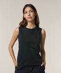 A person with long brown hair stands against a plain light background, wearing the Stanley/Stella Stella Mia (STTW962) sleeveless black top and black trousers, hands in pockets, looking directly at the camera.
