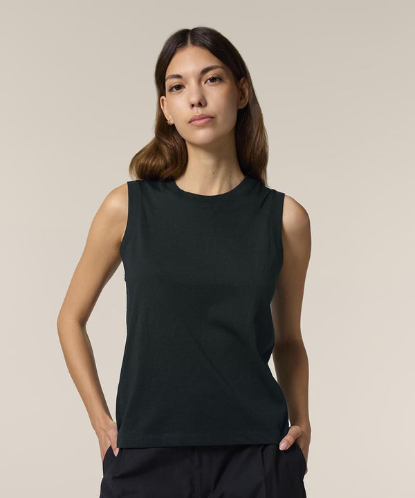 A person with long brown hair stands against a plain light background, wearing the Stanley/Stella Stella Mia (STTW962) sleeveless black top and black trousers, hands in pockets, looking directly at the camera.
