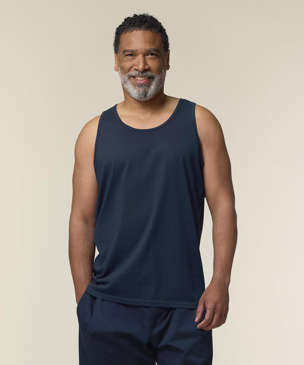 A man with short curly hair and a grey beard smiles whilst standing against a plain light background, wearing the Stanley/Stella Stanley Stancer (STTM961) dark sleeveless vest and dark trousers.