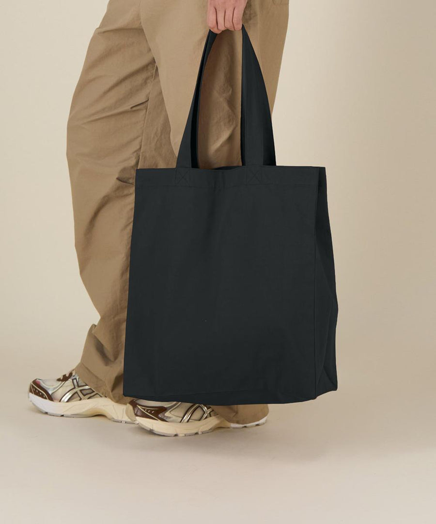 A person in tan trousers and patterned trainers holds the Stanley/Stella Mid tote bag (STAU116) by its handles. The tote bag is large, plain black, and the background is off-white and minimal.
