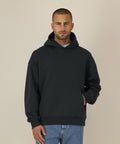 A man with short hair and a trimmed beard wears the Stanley/Stella Chaser (STSU077) black hooded sweatshirt and blue jeans, standing against a beige background with one hand in the front pocket.