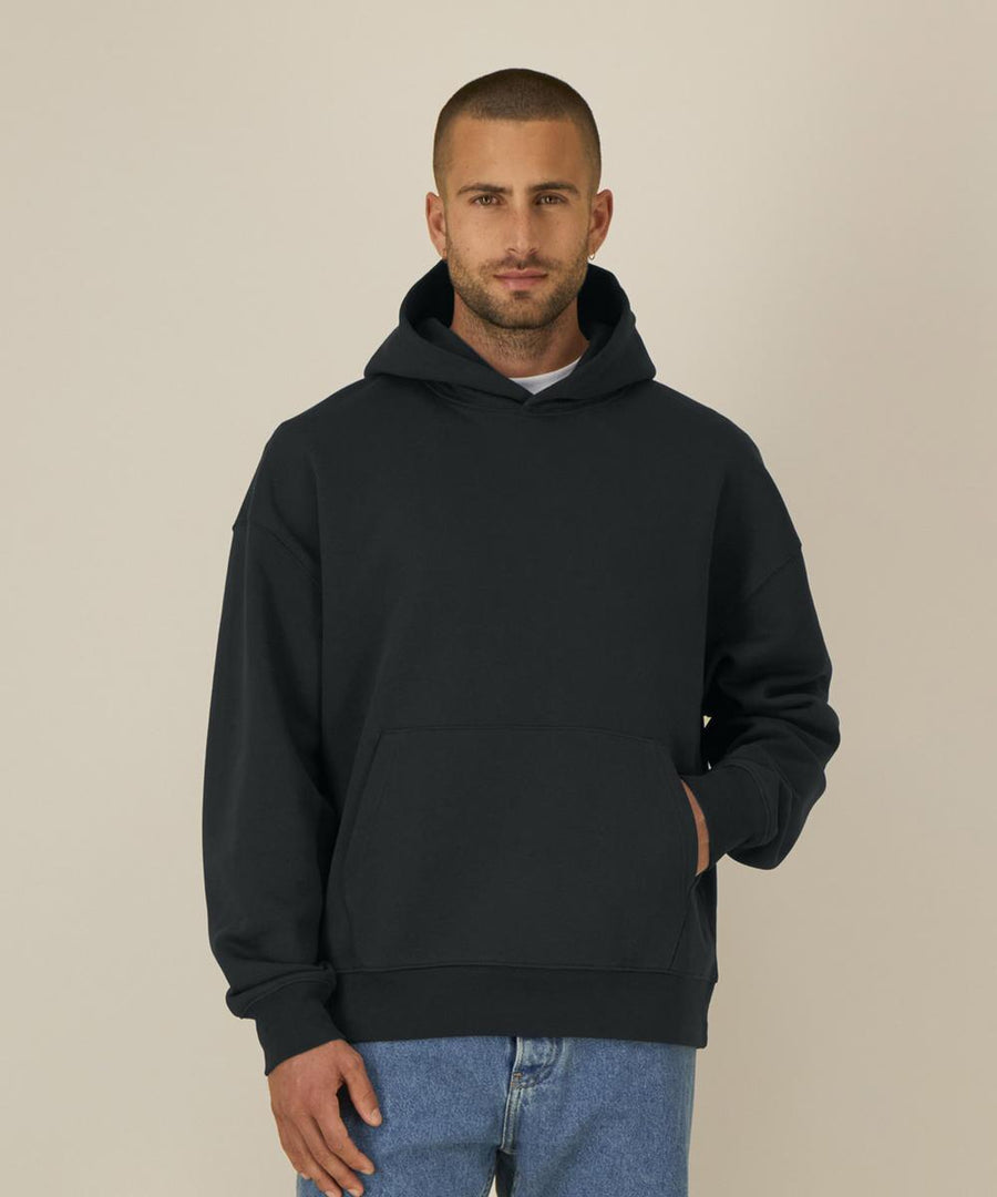 A man with short hair and a trimmed beard wears the Stanley/Stella Chaser (STSU077) black hooded sweatshirt and blue jeans, standing against a beige background with one hand in the front pocket.