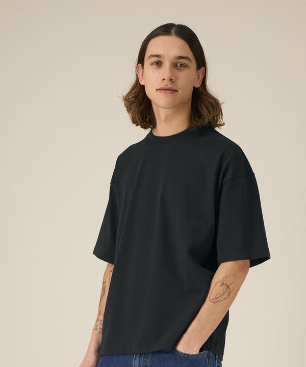 A person with shoulder-length brown hair and tattoos on their forearms wears the Stanley/Stella Asher (STTU078) dark t-shirt and blue jeans, standing against a plain beige background and looking at the camera with a neutral expression.