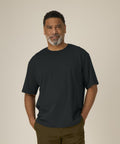 A middle-aged man with short curly hair and a grey beard stands against a beige background, wearing the Stanley/Stella Breezer (STTU964) dark t-shirt and brown trousers, hands in his pockets, smiling slightly.