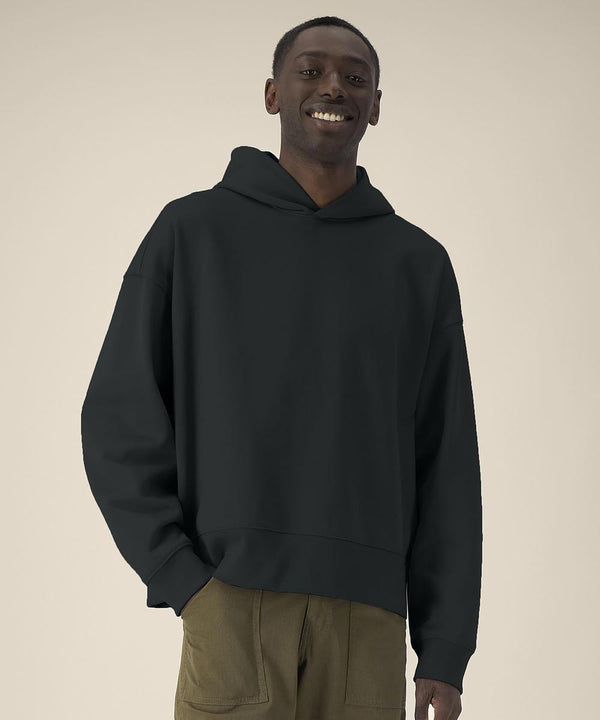 A person stands against a plain beige background, smiling with one hand in their pocket, wearing the Stanley/Stella Voicer (STSU256) black hooded sweatshirt and olive green trousers.