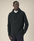 A man stands against a plain beige background wearing the Stanley/Stella Gamechanger (STSU257) dark hooded sweatshirt over a white collared shirt and dark trousers, one hand in his pocket, looking slightly to the side with a neutral expression.