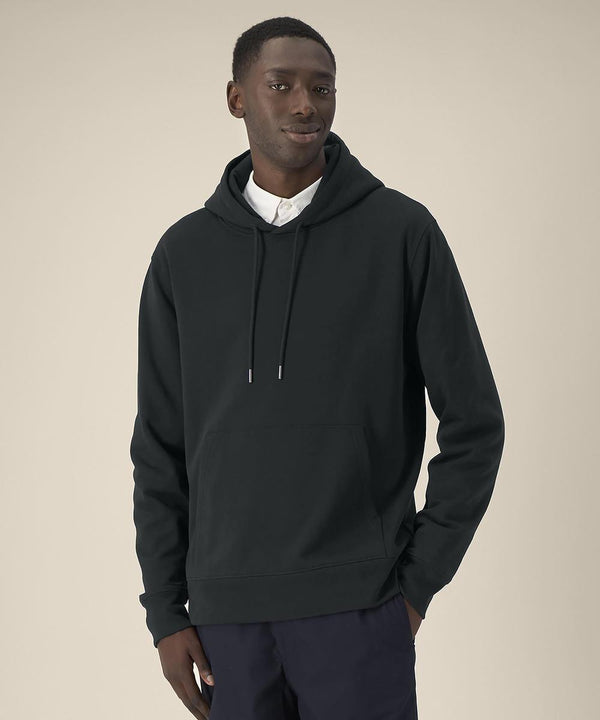 A man stands against a plain beige background wearing the Stanley/Stella Gamechanger (STSU257) dark hooded sweatshirt over a white collared shirt and dark trousers, one hand in his pocket, looking slightly to the side with a neutral expression.