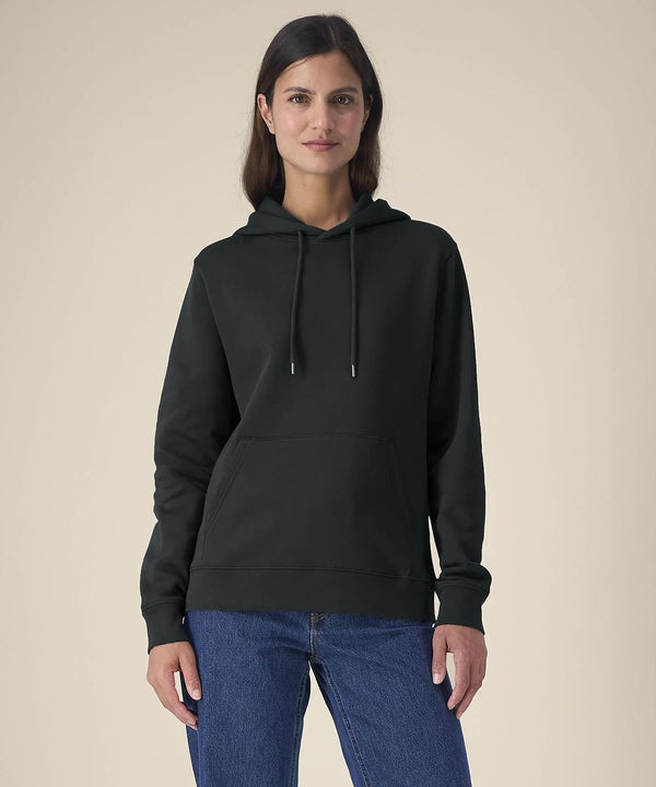 A person with long brown hair stands against a beige background, wearing the black Stanley/Stella Stella Gamechanger (STSW268) hooded sweatshirt and blue jeans, looking directly at the camera with a neutral expression.