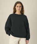 A person with long brown hair models the Stanley/Stella Stella Lianna (STSW262) black sweatshirt and beige trousers, standing against a plain beige background and facing the camera.