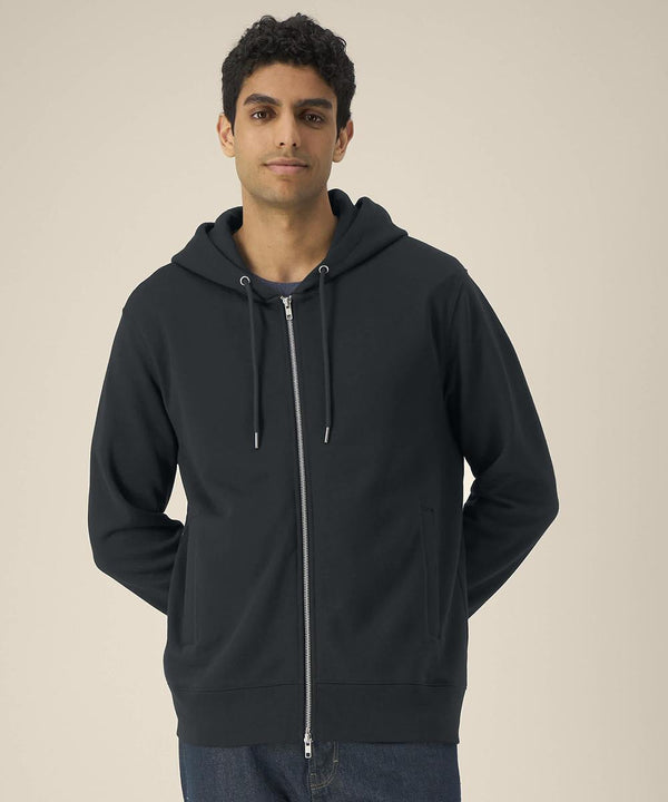 A person faces the camera with a neutral expression, hands behind their back, wearing a Stanley/Stella Astor (STSU261) dark zip-up hoodie with drawstrings and blue jeans, set against a plain light background.