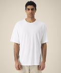 A person with short curly hair wears the Stanley/Stella Blaker (STTU263) plain white oversized T-shirt and light shorts, standing against a beige background and looking straight ahead with a neutral expression.