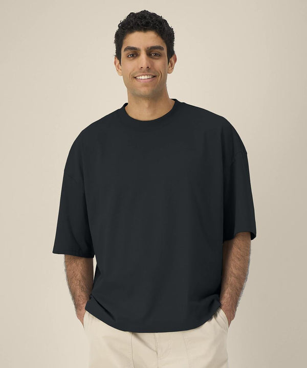 A man with short curly hair stands against a plain beige background, wearing the loose-fitting black Stanley/Stella Hypster (STTU258) t-shirt and light-coloured trousers with his hands in his pockets.