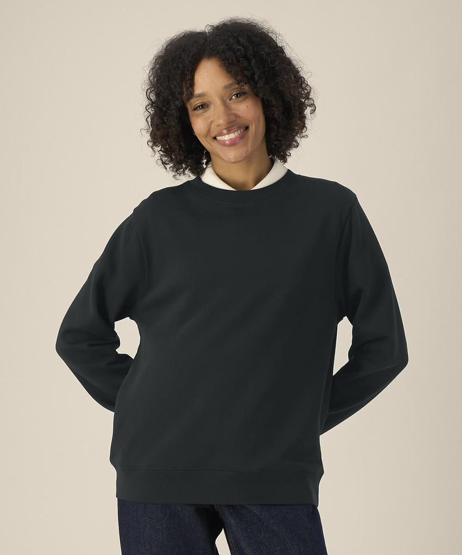 A person with short curly hair wears the Stanley/Stella Stella Thinker (STSW254) dark crew neck sweatshirt over a white collared shirt and blue jeans, standing against a light background and smiling at the camera.