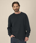 A man with brown hair and a beard wears the Stanley/Stella Thinker (STSU269) plain black long-sleeve sweatshirt and blue jeans, standing against a neutral beige background with one hand in his pocket.