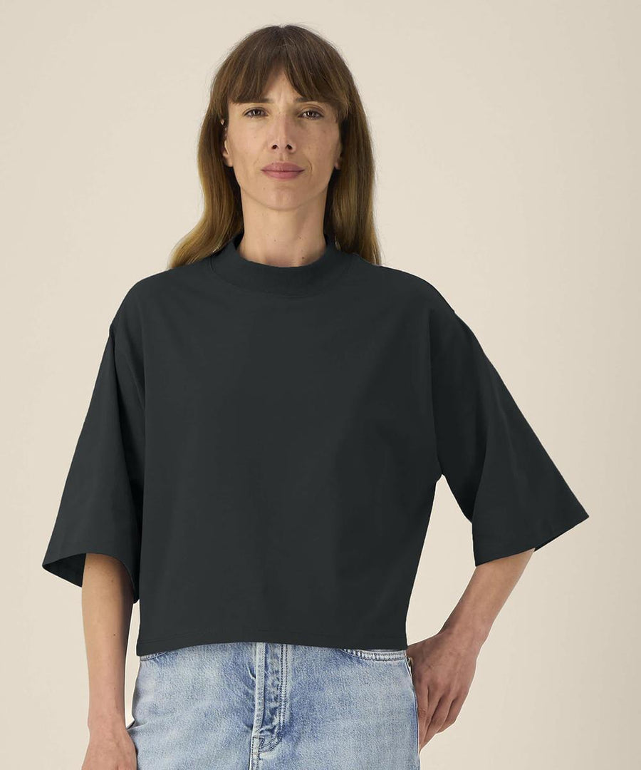 A person with straight, light brown hair models the Stanley/Stella Stella Raya (STTW259) loose black T-shirt with light blue jeans against a plain beige background, standing with a neutral expression and one hand on their hip.