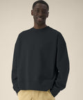 A man stands against a plain beige background, wearing the loose-fitting black Ryler (STSU255) sweatshirt by Stanley/Stella and light tan trousers, with his hands in his pockets and a neutral expression.