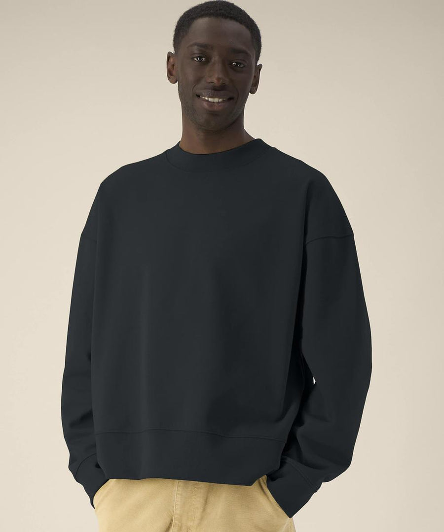 A man stands against a plain beige background, wearing the loose-fitting black Ryler (STSU255) sweatshirt by Stanley/Stella and light tan trousers, with his hands in his pockets and a neutral expression.