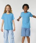 Two children standing against a plain background, both wearing Stanley/Stella SX702 blue t-shirts and denim bottoms. The child on the left has long hair and pockets on the pants, while the child on the right has short hair and shorts.