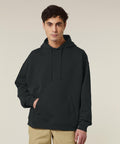 A person with short dark hair stands against a plain background, wearing the Stanley/Stella Slammer 2.0 (STSU209) dark hooded sweatshirt and beige trousers, with one hand in the hoodie's pocket.