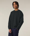 Someone with medium-length twisted hair wears the Stanley/Stella Radder 2.0 (STSU208) black sweatshirt and dark trousers, standing with one hand in their pocket against a neutral background.