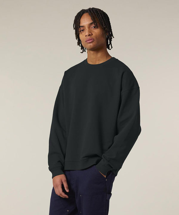 Someone with medium-length twisted hair wears the Stanley/Stella Radder 2.0 (STSU208) black sweatshirt and dark trousers, standing with one hand in their pocket against a neutral background.