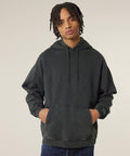 Wearing the Slammer 2.0 Vintage (STSU211) by Stanley/Stella, a person with medium-length dreadlocks stands against a light background in a dark grey hooded sweatshirt and blue jeans, hands in the front pocket.